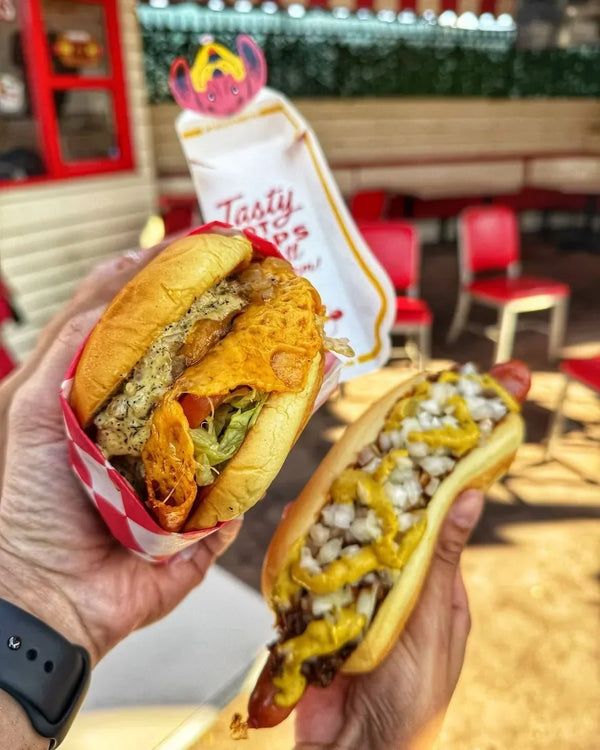 Hands holding a Sassy Cheese Burger and a chili dog topped with onions and yellow mustard at Tail O’ The Pup.
