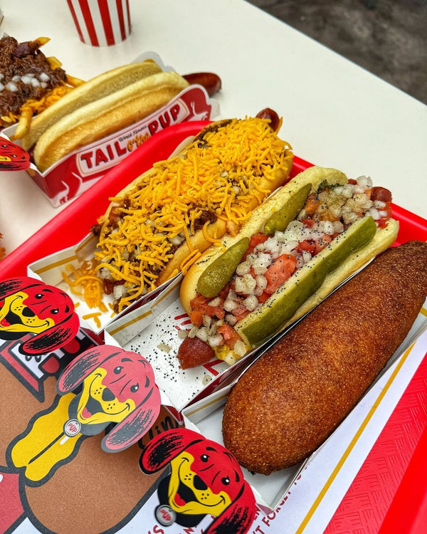 Red tray featuring Tail O’ The Pup’s corn dog, chili cheese dog, and Chicago-style hot dog with pickles and classic toppings.