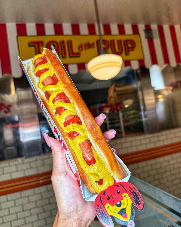 A woman holding a long Tail O’ The Pup hot dog with mustard and ketchup in front of the shop sign.