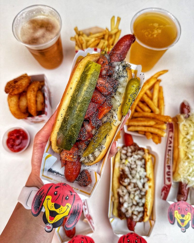A hand holding a loaded Chicago-style hot dog over a table with fries, onion rings, and drinks at Tail O’ The Pup.