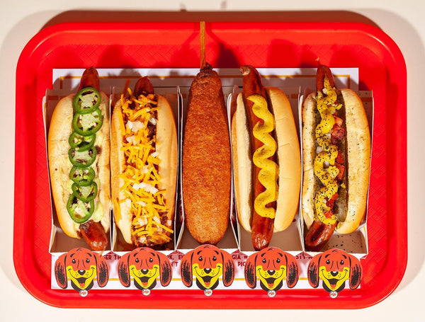 Tail O’ The Pup’s hot dogs with jalapenos, chili cheese, mustard, and relish, and a corn dog, on a red tray.
