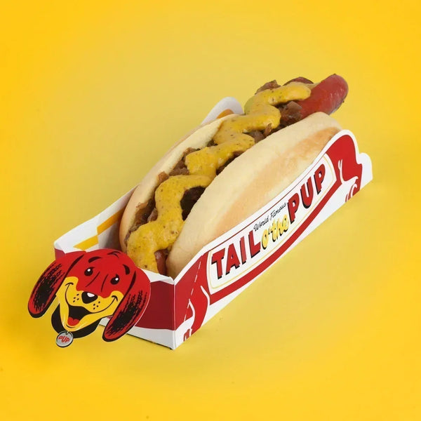 Tail O’ The Pup’s plant-based 1946 hot dog topped with grilled onions and yellow mustard on a yellow backdrop.