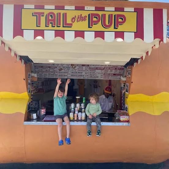 Two children smiling and sitting inside the oversized hot-dog-shaped Tail O’ The Pup stand, enjoying a playful family outing in West Hollywood.