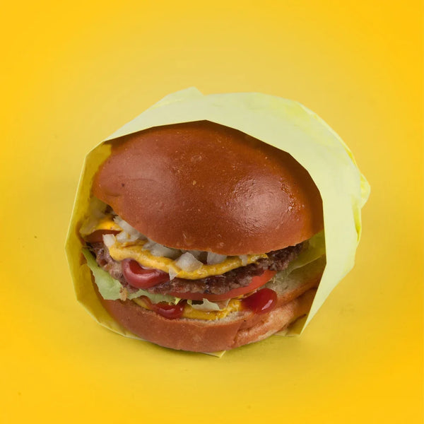 Tail O' The Pup vegan burger wrapped in branded paper with a plant-based patty, onions, mustard, and ketchup on a bright yellow background.