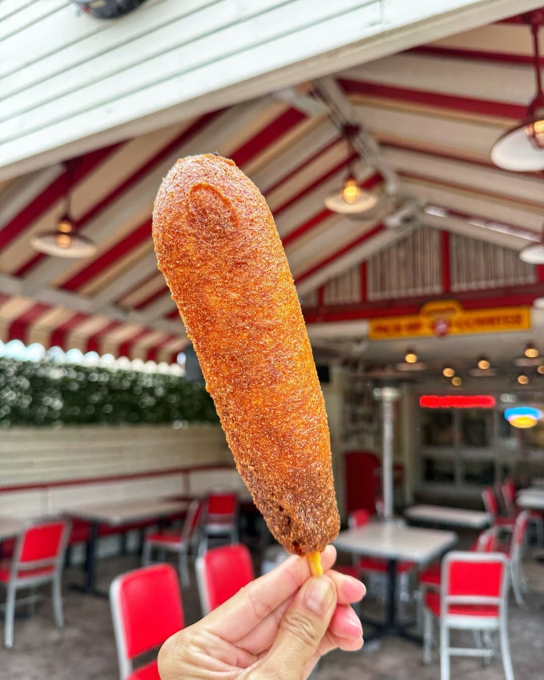 Hand holding a large gluten-free corn dog on a stick in front of the Tail O' The Pup outdoor patio in West Hollywood.
