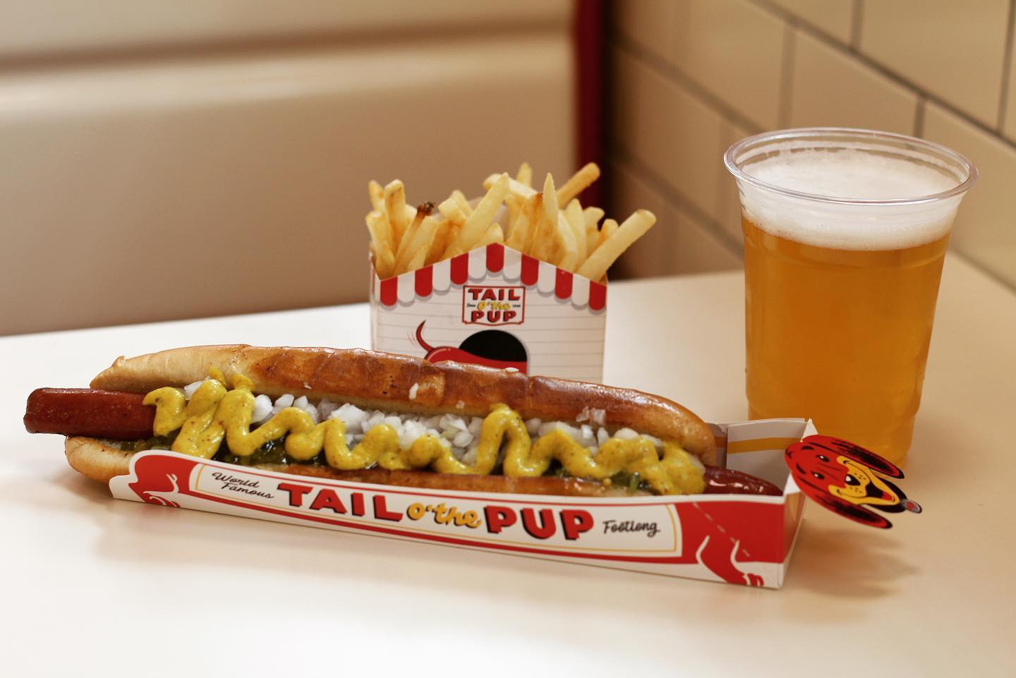 A footlong hot dog with mustard and onions, served with French fries and a glass of beer at Tail O’ The Pup.