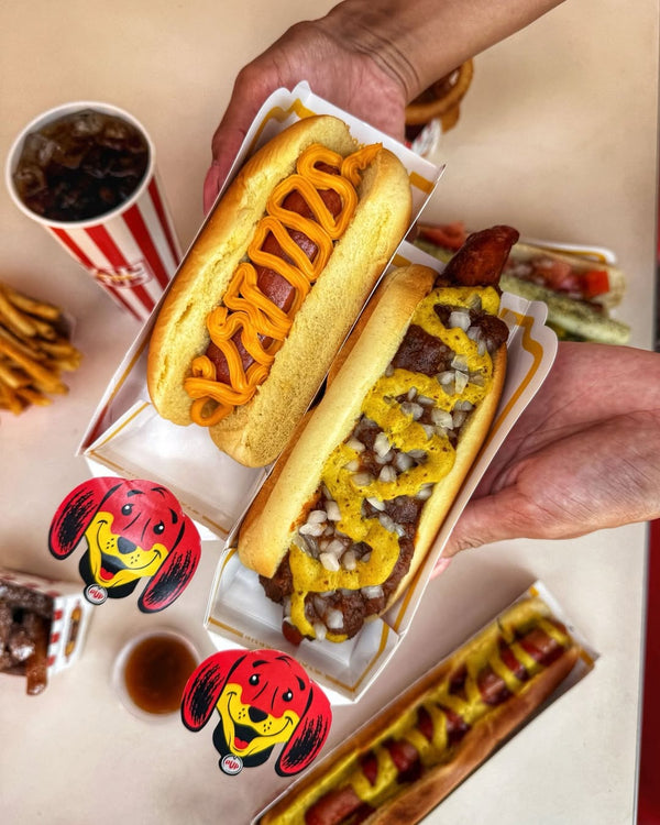 Two hands holding gourmet hot dogs above a table decorated with Tail O' The Pup mascot stickers.