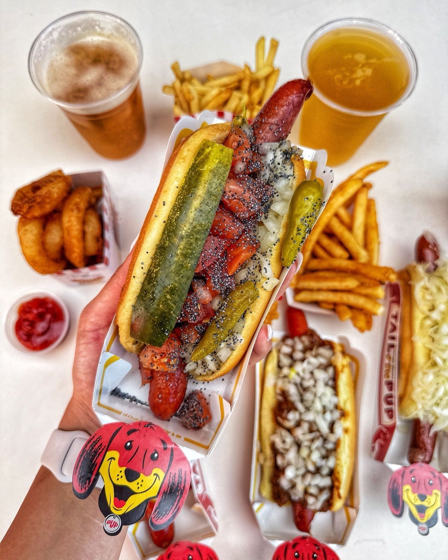 A hand holding a loaded Chicago-style hot dog over a table with fries, onion rings, and drinks at Tail O’ The Pup.