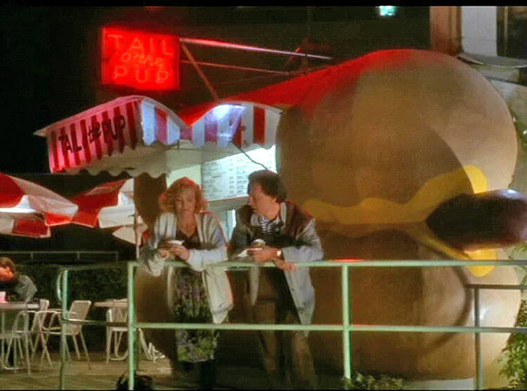 Victoria Tennant and Steve Martin standing beside the neon-lit Tail O’ The Pup hot dog stand at night in Hollywood.