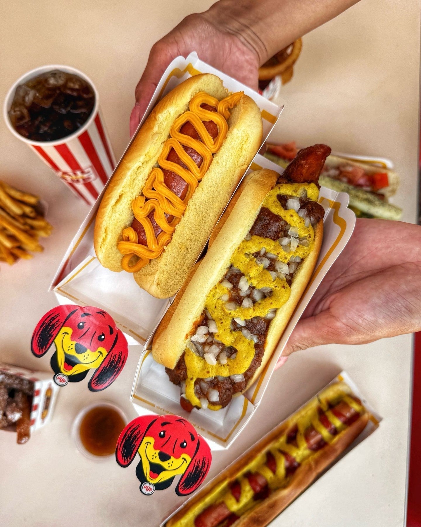 Three Tail O’ The Pup hot dogs with fries and drinks in the background.