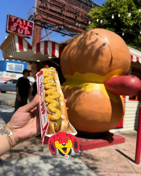 Hand holding a fully loaded hot dog in front of the iconic hot-dog-shaped Tail O' The Pup stand in West Hollywood.